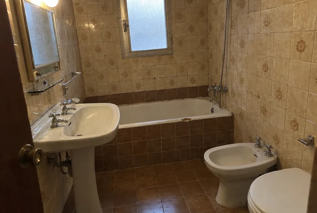 Baño antiguo de un piso de los años setenta con azulejo beige de flores, bañera esmaltada y lavabo de pie
